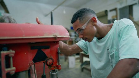Téléchargez les photos : Un jeune apprenti charpentier noir heureux utilisant une machine industrielle se tourne vers la caméra en souriant. Portrait amical d'un travailleur occupant un emploi - en image libre de droit