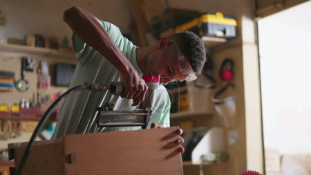 Foto de Un joven carpintero negro construyendo y fijando muebles en el taller de carpintería. Ocupación laboral del aprendiz aprendiendo a construir objetos de madera, utilizando equipos de perforación - Imagen libre de derechos