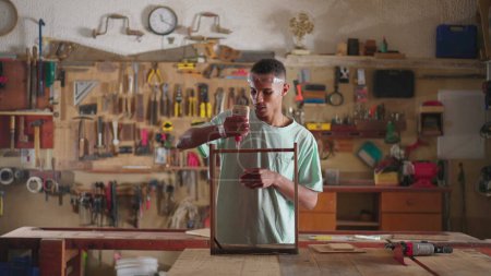 Foto de Un joven carpintero negro dedicado al trabajo en el lugar de trabajo de carpintería, aplicando pegamento en muebles de superficie de madera y pegando un pedazo de madera - Imagen libre de derechos