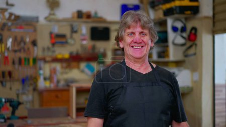 Photo for One joyful master carpenter portrait smiling at camera, wearing apron standing inside carpentry workshop with tools and equipment in background hanging on the wall - Royalty Free Image