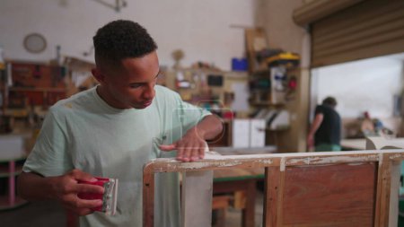 Téléchargez les photos : Un jeune menuisier noir polissant des meubles en bois debout à l'intérieur atelier de menuiserie. Travailleur concentré réparation et création d'objets faits à la main - en image libre de droit