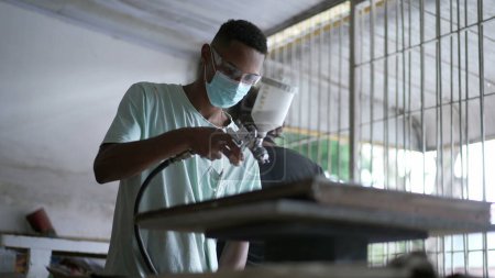 Téléchargez les photos : Un jeune travailleur noir applique un jet de peinture sur une surface en bois dans un atelier de menuiserie - en image libre de droit