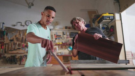Téléchargez les photos : Maître charpentier travaillant à l'atelier de menuiserie avec son jeune apprenti, montrant comment scier le bois à la machine. Jeune homme penché avec l'aide d'un mentor - en image libre de droit