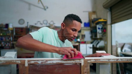Foto de Un joven carpintero brasileño negro puliendo la superficie de madera en el taller de carpintería con herramienta. Empleado comprometido con la ocupación laboral, centrado en la reparación de muebles - Imagen libre de derechos