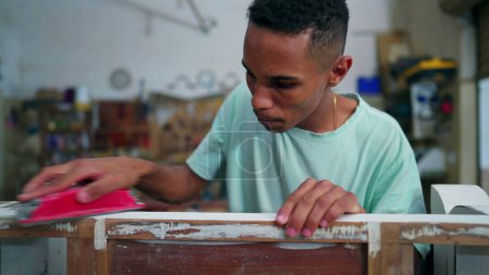 Photo for One young black Brazilian carpenter polishing wooden surface at carpentry workshop with tool. Employee engaged with job occupation, focused in repairing furniture - Royalty Free Image
