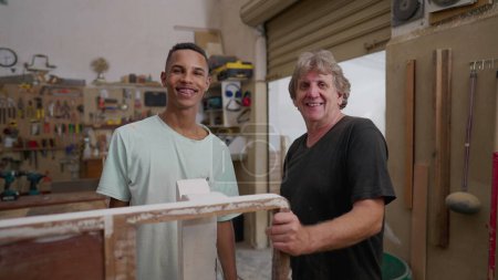 Téléchargez les photos : Portrait de jeunes apprentis charpentiers souriants à la caméra debout à l'intérieur de l'atelier de menuiserie. Authentiques personnes de la vie réelle, profession - en image libre de droit