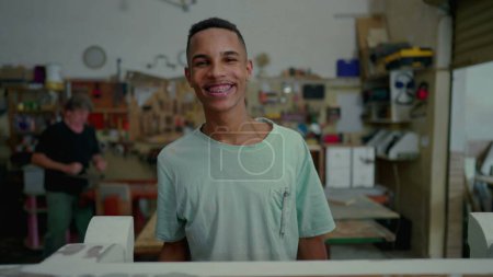 Foto de Retrato de un empleado brasileño negro feliz de taller de carpintería, ocupación local de pequeñas empresas. Joven aprendiz mirando a la cámara sonriendo - Imagen libre de derechos