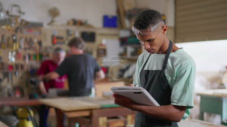 Photo for One young black apprentice of carpentry workshop taking notes on notepad, wearing apron and protective eye-wear. Staff employee engaged in learning business - Royalty Free Image
