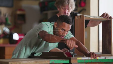 Téléchargez les photos : Un jeune élève noir mesurant le bois avec règle à l'atelier, Maître charpentier prenant des notes, Scène franche de charpentiers travaillant - en image libre de droit