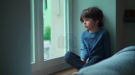 Téléchargez les photos : Enfant ennuyé assis à l'intérieur par la fenêtre de l'appartement regardant du deuxième étage voulant aller à l'extérieur, petit garçon avec une émotion triste - en image libre de droit