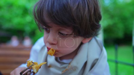 Foto de Niño pequeño y desordenado comiendo pasta para el almuerzo en un entorno soleado al aire libre. Rostro retrato de cerca de fideos saborizantes para niños - Imagen libre de derechos