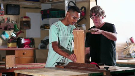 Photo for Candid senior carpenter guiding apprentice to use saw machine, slicing piece of wood at workshop - Royalty Free Image