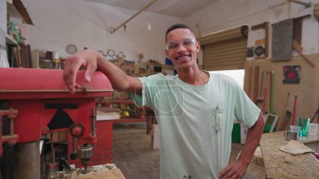 Téléchargez les photos : Un jeune travailleur brésilien noir debout à côté d'une machine industrielle à l'intérieur de l'atelier, portant des lunettes de protection souriant à la caméra avec une expression joyeuse - en image libre de droit