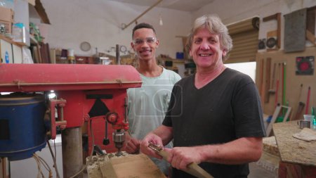 Foto de Retrato de trabajadores brasileños de pie en el interior del taller sonriendo a la cámara. Maestro carpintero sudamericano junto a su joven aprendiz, mirando a la cámara - Imagen libre de derechos