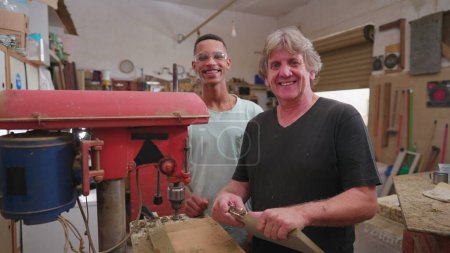 Photo for Portrait of Brazilian workers standing inside workshop smiling at camera. South American master carpenter next to his young apprentice, looking at camera - Royalty Free Image