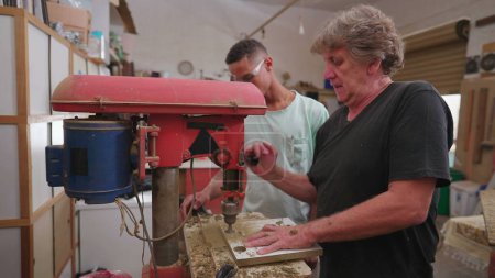 Téléchargez les photos : Maître charpentier utilisant une machine industrielle pour faire un trou dans la surface en bois tout en enseignant à l'apprenti à utiliser l'équipement, portrait des travailleurs brésiliens - en image libre de droit