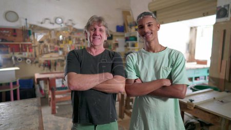 Foto de Carpinteros brasileños de pie en el interior del taller sonriendo a la cámara, cruzando brazos. Retrato de aprendiz y maestro carpintero - Imagen libre de derechos