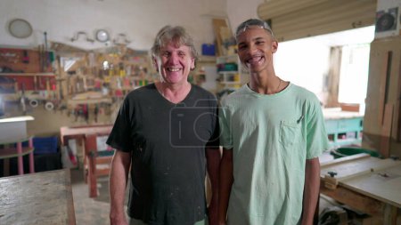 Foto de Carpinteros brasileños de pie en el interior del taller sonriendo a la cámara, cruzando brazos. Retrato de aprendiz y maestro carpintero - Imagen libre de derechos