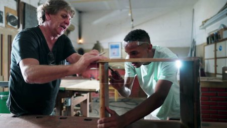 Foto de Maestro carpintero guiando a un joven aprendiz negro en el taller de carpintería, personas comprometidas con su ocupación laboral, trabajo en la industria, trabajando con sus manos. Los brasileños en el trabajo - Imagen libre de derechos