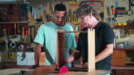 Photo for Master carpenter guiding apprentice at carpentry workshop, boss teaching student with woodwork using saw machine to slice piece of wood, industry job - Royalty Free Image