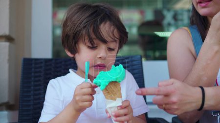 Photo for Child enjoying ice-cream cone at parlo shop during summer day. Mother brings cone closer to small boy's mouth to prevent from staining clothes - Royalty Free Image