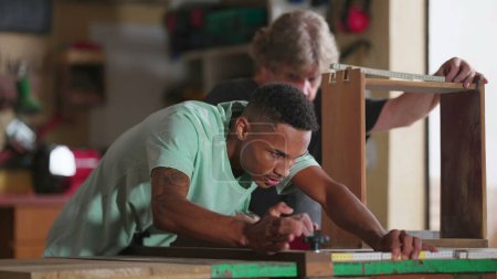 Foto de Un joven alumno negro midiendo madera con gobernante en el taller, maestro carpintero tomando notas, escena cándida de carpinteros trabajando - Imagen libre de derechos