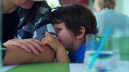 Téléchargez les photos : Petit garçon ennuyé appuyé sur le bras de la mère se sentant l'ennui au restaurant en attendant que la nourriture arrive. Enfant attaché à sa mère grimaçant et frottant le visage - en image libre de droit