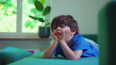 Foto de Niño pequeño viendo dibujos animados fuera de cámara con la mano en la barbilla absorbida por el contenido de entretenimiento colocado en el sofá sofá - Imagen libre de derechos