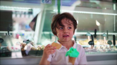 Photo for One small boy holding ice-cream cone taking a bite of biscuit. Child eating colorful green dessert indulgence at ice cream parlor - Royalty Free Image