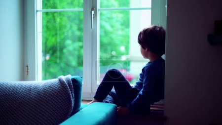 Foto de Pequeño niño reflexivo mirando la lluvia a través de la ventana del apartamento, mirada contemplativa durante el tiempo lluvioso, expresión pensativa del niño - Imagen libre de derechos