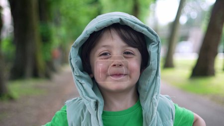 Téléchargez les photos : Portrait d'un beau petit garçon heureux debout dehors tout en portant un sweat à capuche. Gros plan de l'enfant au parc - en image libre de droit