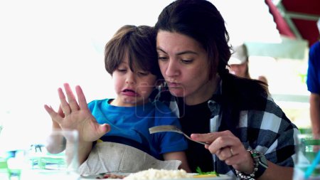 Photo for Mother feeding restless child at restaurant, kid sitting on mom's lap being fed food during lunch time. Family traveling on vacation eating out, candid and authentic - Royalty Free Image