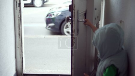 Téléchargez les photos : Un enfant ouvre la porte de la maison et sort dans la rue. La main d'un petit garçon atteint le bouton de la porte et ouvre l'entrée de la résidence tout en portant un sweat à capuche - en image libre de droit
