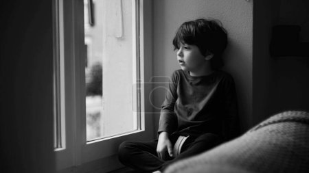 Photo for Sad bored child sitting by window looking out with pensive contemplative expression in monochromatic, black and white - Royalty Free Image