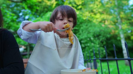 Téléchargez les photos : Nouilles pour enfants savourant des pâtes pour le déjeuner en plein air, petit col garçon recouvert de serviette pour éviter les taches, enfant mangeant des glucides par lui-même - en image libre de droit
