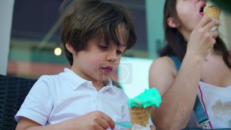 Téléchargez les photos : Enfant mangeant cône coloré de crème glacée avec maman à l'extérieur. Un petit garçon caucasien apprécie le dessert sucré pendant la journée d'été - en image libre de droit