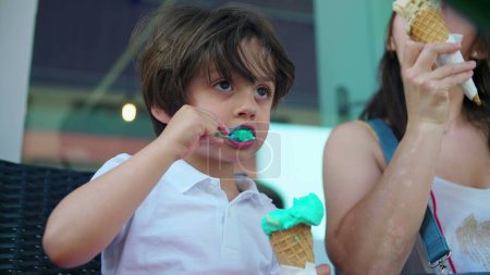 Foto de Niño comiendo cono de helado colorido con mamá afuera. Un niño caucásico disfruta de postre dulce durante el día de verano - Imagen libre de derechos