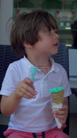 Foto de Niño pequeño disfrutando del cono de helado con cuchara. Niño comiendo colorido helado azul, dulce postre comida de verano, vídeo vertical - Imagen libre de derechos