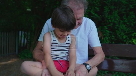 Photo for Loving family moment of grandfather bonding with grandson seated at park bench during summer day. Authentic loving affectionate moment between inter-generational people - Royalty Free Image