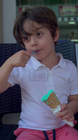 Photo for Small boy enjoying ice-cream cone with spoon. Child eating colorful blue icecream, treat summer food dessert, vertical video - Royalty Free Image