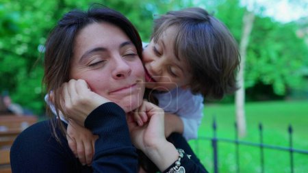 Foto de Momento de maternidad de niño colgando de la espalda de mamá al aire libre afuera, besando y abrazando a mamá con amor y afecto auténtico. Cuidar niño pequeño abraza y besa a los padres - Imagen libre de derechos