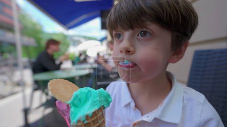 Foto de Little Boy saborea el cono de helado en un día de verano. Niño lamiendo colorido helado azul afuera - Imagen libre de derechos