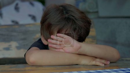 Téléchargez les photos : Enfant ennuyé couvrant le visage avec les bras appuyés sur la table se sentant l'ennui et la tristesse, l'enfant lutte avec l'isolement, rien à faire - en image libre de droit