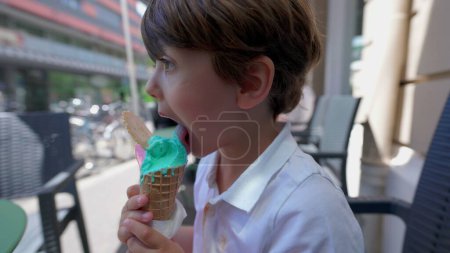 Photo for Small boy eating colorful icecream cone outside at parlor shop's sidewalk. Profile face of kid licking sweet dessert indulgence - Royalty Free Image