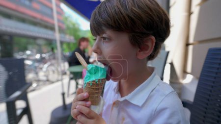 Téléchargez les photos : Petit garçon mangeant cône de glace coloré à l'extérieur sur le trottoir du magasin de salon. Profil visage d'enfant léchant doux dessert indulgence - en image libre de droit