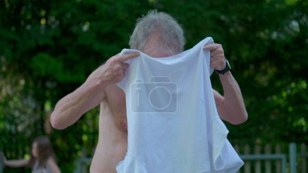 Téléchargez les photos : Homme âgé mettant t-shirt dehors dans la journée ensoleillée. Vêtements de personne âgée lui-même pendant la journée d'été - en image libre de droit