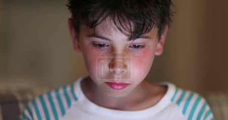 Foto de Niño delante de la pantalla de la tableta con luz azul que brilla en la cara por la noche. Niño viendo contenido en línea navegar por Internet - Imagen libre de derechos