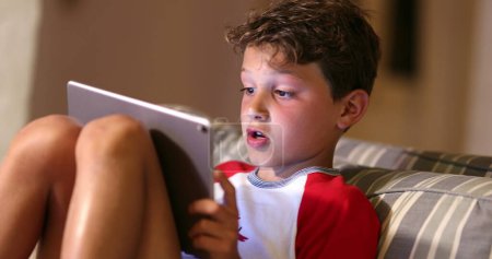 Téléchargez les photos : Boy playing game with tablet. Digital native child plays video-game on tech device at night. Kid surprise emotion - en image libre de droit