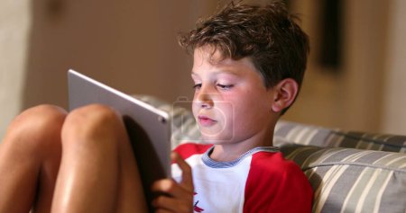 Foto de Boy playing game with tablet. Digital native child plays video-game on tech device at night. Kid surprise emotion - Imagen libre de derechos