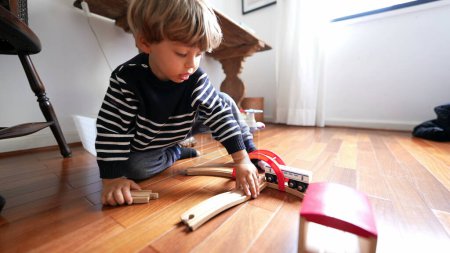 Téléchargez les photos : Enfant joue avec des jouets rétro sur le plancher de bois franc. Un petit garçon jouant à l'intérieur avec des rails en bois de train par lui-même - en image libre de droit
